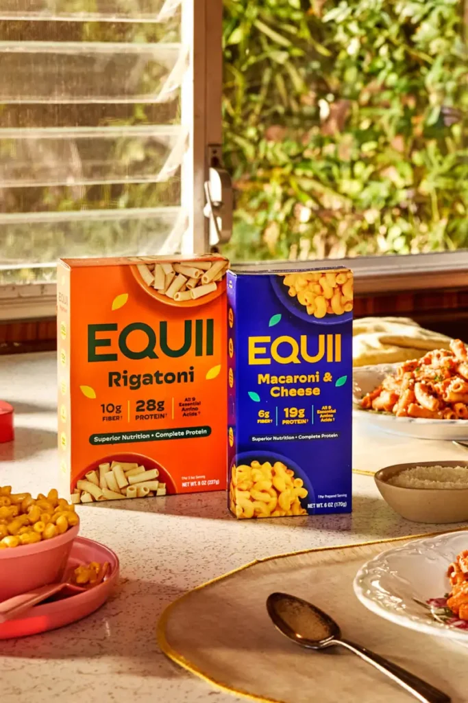 Boxes of EQUII Rigatoni and Macaroni & Cheese on a kitchen table, surrounded by pasta dishes and greenery outside.