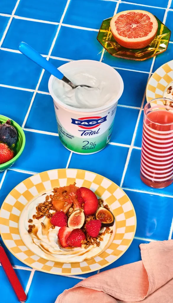 Yogurt with figs, strawberries, granola on a plate, next to a yogurt tub, half grapefruit, and juice on a blue tiled table.