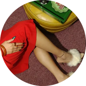 Woman in a red dress with green nails and fluffy heels, sitting next to a round table on a burgundy carpet.
