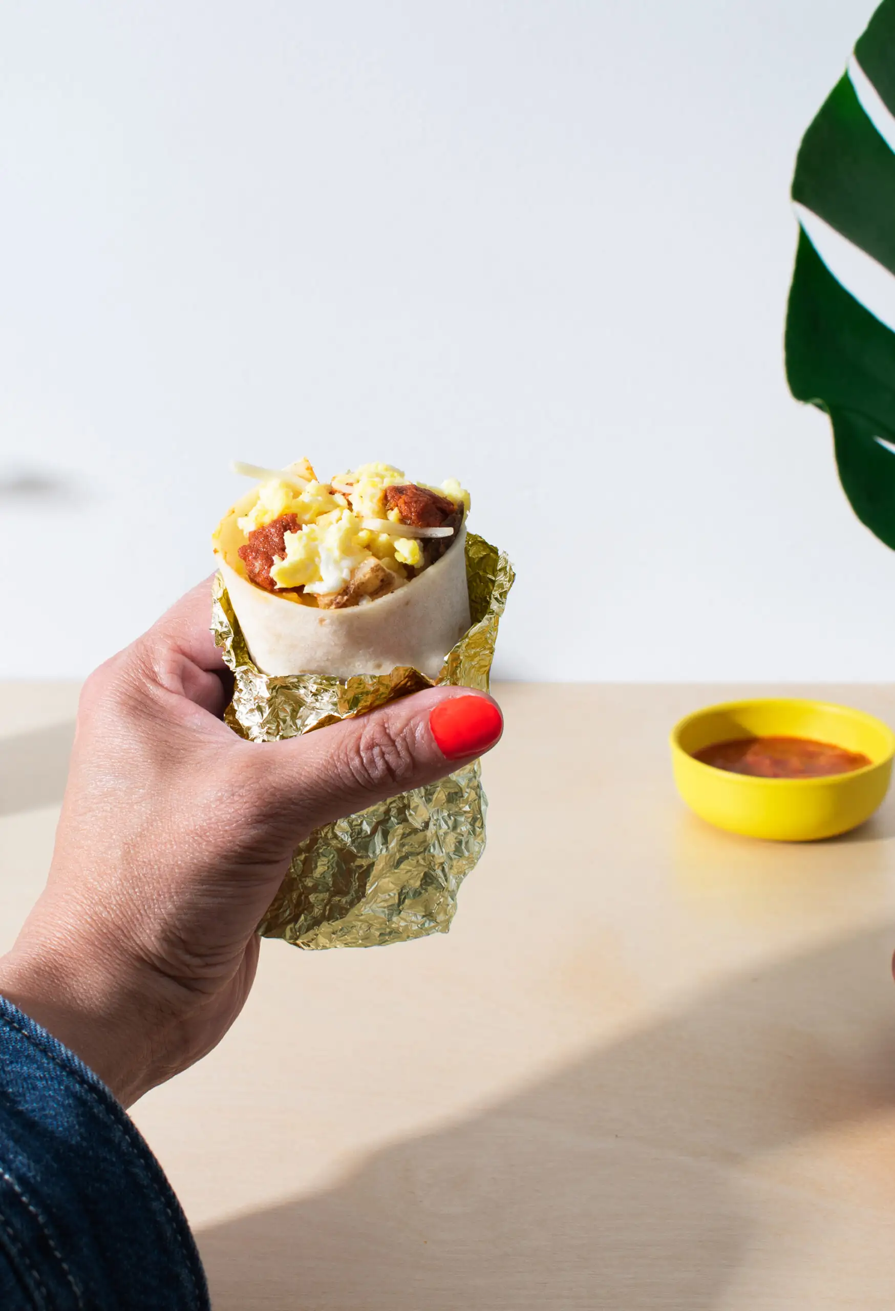 Hand holding a breakfast burrito wrapped in foil, with a yellow bowl of sauce and a green plant in the background.