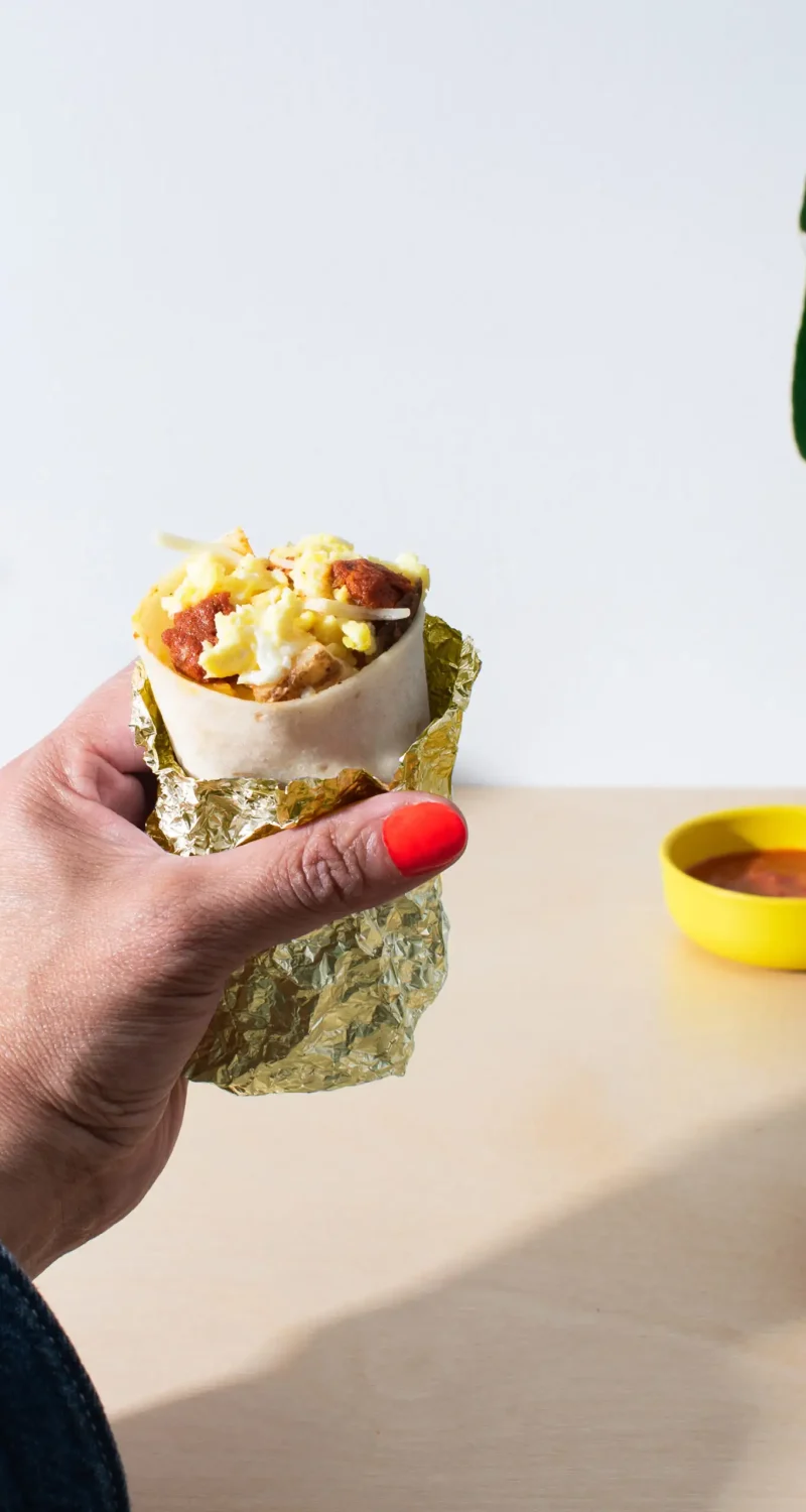 Hand holding a breakfast burrito with eggs and chorizo, wrapped in foil, with a yellow bowl of sauce on the table.