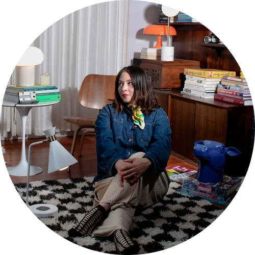 Woman in denim jacket sitting on patterned rug, surrounded by books and decor, with colorful lighting in the background.