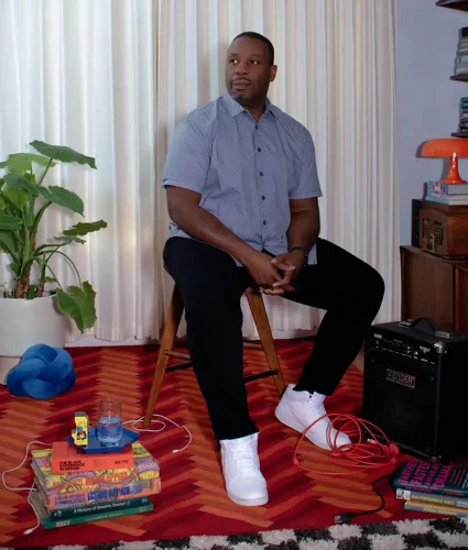 Man seated on a chair in a room with a patterned rug, surrounded by books, a plant, an amplifier, and a small blue sculpture.