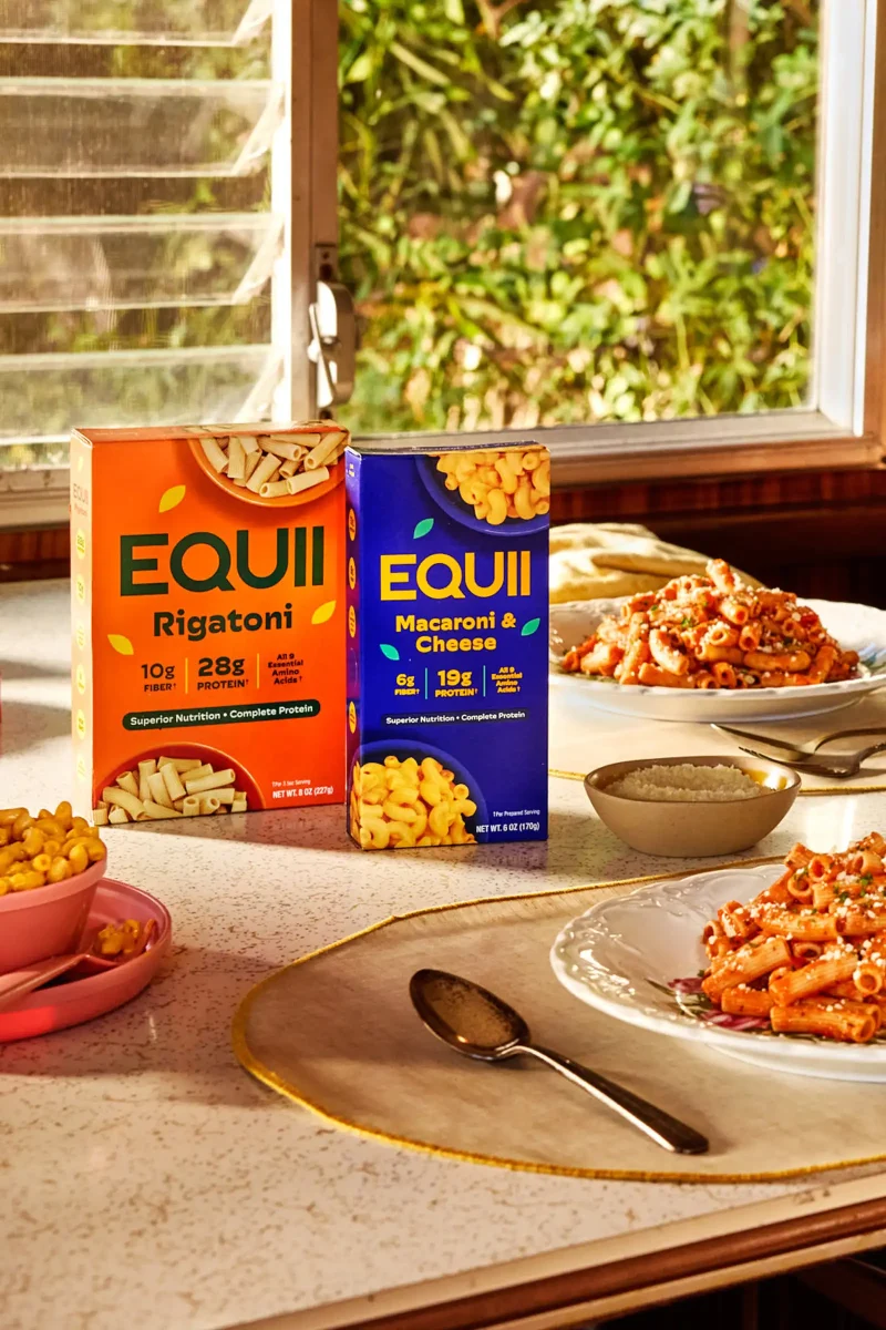 Boxes of Equii Rigatoni and Macaroni & Cheese on a table with prepared dishes by a sunlit window.