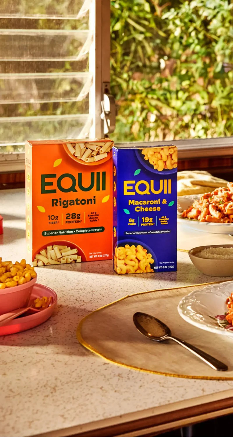 Boxes of EQUII Rigatoni and Macaroni & Cheese on a table, surrounded by bowls and plates, with a garden view.