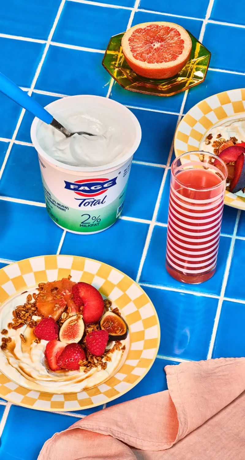 Breakfast setting on blue tiles with yogurt, granola, fruit, grapefruit, and a striped glass of juice.